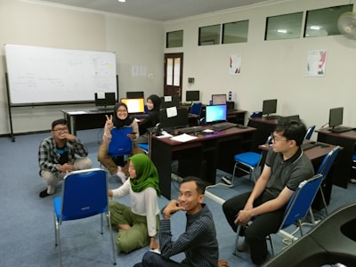 A group of six people are gathered in a computer room, some sitting on the floor and others on chairs. They are casually dressed and appear to be engaged in a relaxed activity, with one person flashing a peace sign and another smiling brightly. The room contains several computers on desks and a large whiteboard in the background with some writing on it.