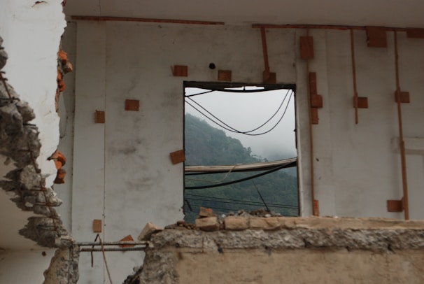 A partially demolished building with exposed concrete and rebar, featuring a window frame with a view of misty hills in the background. The wall has wooden reinforcements and appears weathered, creating a contrast between the indoor destruction and the serene outdoor landscape.