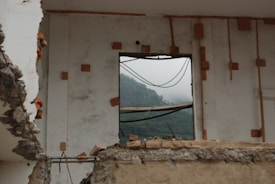 A partially demolished building with exposed concrete and rebar, featuring a window frame with a view of misty hills in the background. The wall has wooden reinforcements and appears weathered, creating a contrast between the indoor destruction and the serene outdoor landscape.