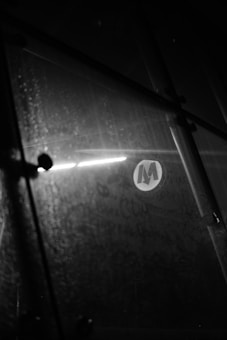 A black and white photograph featuring a glass surface with droplets and smudges, along with geometric patterns. A circular logo with the letter 'M' is visible near a bright reflective light source, creating a stark contrast.