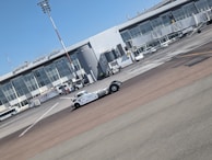 A vintage-style white vehicle is parked on the tarmac in front of a modern airport terminal building with large glass windows. Several ground support vehicles and equipment are also present. The sky is clear and blue.