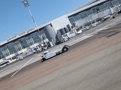 A vintage-style white vehicle is parked on the tarmac in front of a modern airport terminal building with large glass windows. Several ground support vehicles and equipment are also present. The sky is clear and blue.