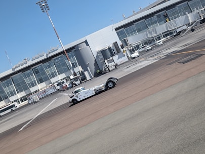 A vintage-style white vehicle is parked on the tarmac in front of a modern airport terminal building with large glass windows. Several ground support vehicles and equipment are also present. The sky is clear and blue.