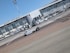 A vintage-style white vehicle is parked on the tarmac in front of a modern airport terminal building with large glass windows. Several ground support vehicles and equipment are also present. The sky is clear and blue.