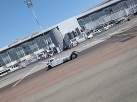 A vintage-style white vehicle is parked on the tarmac in front of a modern airport terminal building with large glass windows. Several ground support vehicles and equipment are also present. The sky is clear and blue.