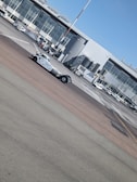 A busy airport tarmac featuring a range of vehicles including a tow truck in the foreground. The backdrop includes a terminal building with large glass windows and multiple jet bridges. Several small airport vehicles and equipment are scattered around the area.
