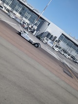 A busy airport tarmac featuring a range of vehicles including a tow truck in the foreground. The backdrop includes a terminal building with large glass windows and multiple jet bridges. Several small airport vehicles and equipment are scattered around the area.