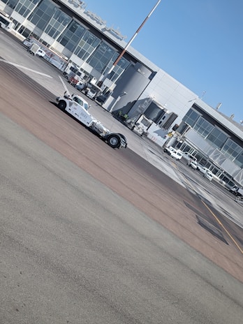A busy airport tarmac featuring a range of vehicles including a tow truck in the foreground. The backdrop includes a terminal building with large glass windows and multiple jet bridges. Several small airport vehicles and equipment are scattered around the area.