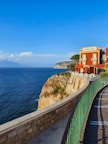 A vibrant red building labeled 'Albergo Lorelei' stands majestically on a cliff overlooking a vast blue ocean. The structure features ornate windows and a terrace with tables and chairs. A stone pathway with a green railing curves gracefully along the edge of the cliff. In the distance, gentle waves meet the rocky landscape and a hazy mountain range under a clear blue sky adorned with fluffy clouds.