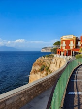 A vibrant red building labeled 'Albergo Lorelei' stands majestically on a cliff overlooking a vast blue ocean. The structure features ornate windows and a terrace with tables and chairs. A stone pathway with a green railing curves gracefully along the edge of the cliff. In the distance, gentle waves meet the rocky landscape and a hazy mountain range under a clear blue sky adorned with fluffy clouds.