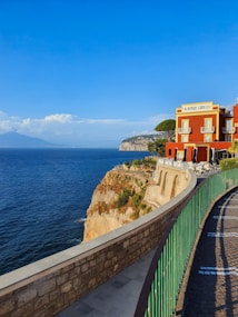A vibrant red building labeled 'Albergo Lorelei' stands majestically on a cliff overlooking a vast blue ocean. The structure features ornate windows and a terrace with tables and chairs. A stone pathway with a green railing curves gracefully along the edge of the cliff. In the distance, gentle waves meet the rocky landscape and a hazy mountain range under a clear blue sky adorned with fluffy clouds.