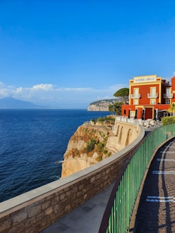 A vibrant red building labeled 'Albergo Lorelei' stands majestically on a cliff overlooking a vast blue ocean. The structure features ornate windows and a terrace with tables and chairs. A stone pathway with a green railing curves gracefully along the edge of the cliff. In the distance, gentle waves meet the rocky landscape and a hazy mountain range under a clear blue sky adorned with fluffy clouds.
