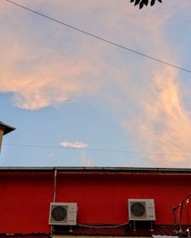 A red building wall is visible with two air conditioning units mounted on it. Above the building, the sky displays soft clouds with hues of orange and pink, suggesting a sunset or sunrise. A thin wire crosses horizontally, and some tree leaves are visible in the corner of the image.