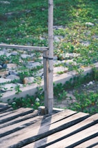 Wooden boards form a small rustic deck or pathway in a green, outdoor setting, featuring a simple bamboo railing tied together with a dark band. The ground below is covered in lush vegetation with small, colorful flowers visible among the leaves.