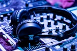 A pair of black headphones is placed on a DJ mixer. The mixer has various knobs and sliders, and the lighting gives off a blue-tinted ambiance, suggesting a musical or party setting.
