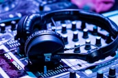 A pair of black headphones is placed on a DJ mixer. The mixer has various knobs and sliders, and the lighting gives off a blue-tinted ambiance, suggesting a musical or party setting.