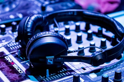 A pair of black headphones is placed on a DJ mixer. The mixer has various knobs and sliders, and the lighting gives off a blue-tinted ambiance, suggesting a musical or party setting.