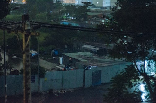 A dimly lit urban area at night, featuring metallic rooftops and a fenced perimeter. There are various structures and possibly construction equipment scattered around. Dark trees frame the scene, with faint city lights visible in the distance.