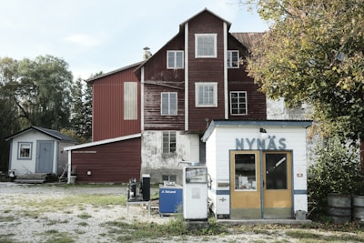 A rustic scene featuring an old-fashioned gas station with the sign 'NYNÄS' on a small white building with yellow doors. Next to the station are vintage fuel pumps labeled 'BRA ADEL' and 'DE LAVAL,' resting beside a larger red wooden building with a weathered look. Surrounding the structures are trees with green leaves, indicating a rural landscape. The setting feels quiet and historical with a touch of nostalgia.