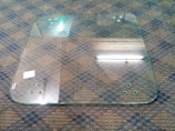 A large, transparent glass panel lies on a checkered carpeted floor. It has circular holes near the corners, and the edges appear slightly rounded. A reflection of fluorescent lights can be seen on the glass surface, and there is a small white sticker or label on one side.