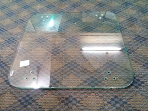 A large, transparent glass panel lies on a checkered carpeted floor. It has circular holes near the corners, and the edges appear slightly rounded. A reflection of fluorescent lights can be seen on the glass surface, and there is a small white sticker or label on one side.