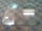 A large, rectangular glass panel with rounded corners lies on a patterned carpeted floor. The glass has six small circular holes near the edges, with some markings and scratches visible on the surface. There's a small white sticker on one corner of the glass panel. Reflections of overhead fluorescent lights can be seen on the glass.