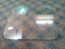 A large, rectangular glass panel with rounded corners lies on a patterned carpeted floor. The glass has six small circular holes near the edges, with some markings and scratches visible on the surface. There's a small white sticker on one corner of the glass panel. Reflections of overhead fluorescent lights can be seen on the glass.