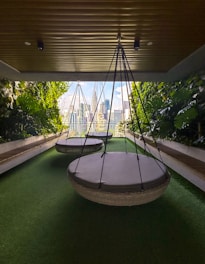 A modern outdoor seating area with large circular hanging chairs suspended from the ceiling. The space is bordered by lush green plants and features synthetic green grass flooring. In the background, a cityscape with tall buildings can be seen through glass walls, allowing natural light to filter in.