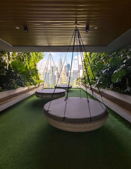A modern outdoor seating area with large circular hanging chairs suspended from the ceiling. The space is bordered by lush green plants and features synthetic green grass flooring. In the background, a cityscape with tall buildings can be seen through glass walls, allowing natural light to filter in.