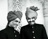 Two men stand side by side, both wearing traditional dark-colored attire with button-up jackets and intricately wrapped turbans. They are smiling, with one showing a more pronounced grin. The background features ornate stonework with floral patterns, suggesting a historical or cultural setting.