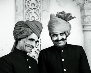 Two men stand side by side, both wearing traditional dark-colored attire with button-up jackets and intricately wrapped turbans. They are smiling, with one showing a more pronounced grin. The background features ornate stonework with floral patterns, suggesting a historical or cultural setting.
