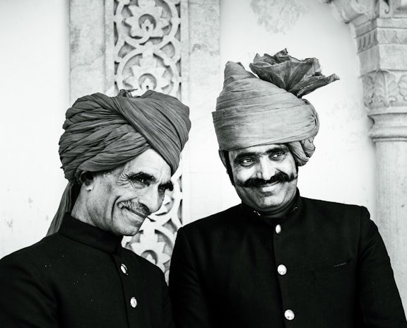 Two men stand side by side, both wearing traditional dark-colored attire with button-up jackets and intricately wrapped turbans. They are smiling, with one showing a more pronounced grin. The background features ornate stonework with floral patterns, suggesting a historical or cultural setting.