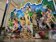 A detailed sculpture depicting multiple Hindu deities, including Shiva, Durga, and a lion. The scene is vibrant and colorful, set against a painted blue sky with clouds and greenery in the background. Various deities are depicted in traditional attire, surrounded by plants and mythical creatures.