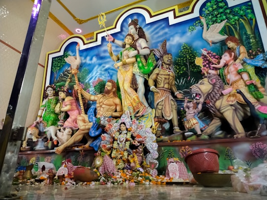 A detailed sculpture depicting multiple Hindu deities, including Shiva, Durga, and a lion. The scene is vibrant and colorful, set against a painted blue sky with clouds and greenery in the background. Various deities are depicted in traditional attire, surrounded by plants and mythical creatures.