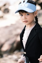 A young child wearing a light blue visor with bunny ears, and a smart black blazer over a simple shirt. The child has braided hair and is looking towards the camera with a calm expression. The background is blurred, suggesting an outdoor setting.