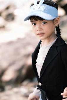 A young child wearing a light blue visor with bunny ears, and a smart black blazer over a simple shirt. The child has braided hair and is looking towards the camera with a calm expression. The background is blurred, suggesting an outdoor setting.