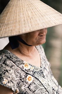 An elderly person wears a traditional conical hat, partially obscuring their face. The individual is dressed in a patterned garment, featuring floral designs. The mood is contemplative, with soft lighting casting gentle shadows.