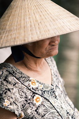 An elderly person wears a traditional conical hat, partially obscuring their face. The individual is dressed in a patterned garment, featuring floral designs. The mood is contemplative, with soft lighting casting gentle shadows.