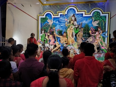 A group of people gather around a colorful and elaborate religious display featuring deities and mythological figures. The scene includes several statues, including divine figures with traditional attire and ornate details. People are dressed in red and other vibrant colors, observing the display in a reverent manner within an interior setting.