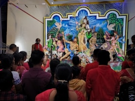 A group of people gather around a colorful and elaborate religious display featuring deities and mythological figures. The scene includes several statues, including divine figures with traditional attire and ornate details. People are dressed in red and other vibrant colors, observing the display in a reverent manner within an interior setting.