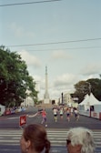 A group of people is participating in a running event on a city street, with a tall monument in the background. Tents and barriers line the street, and trees are visible on the left side. The weather appears partly cloudy.