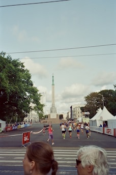 A group of people is participating in a running event on a city street, with a tall monument in the background. Tents and barriers line the street, and trees are visible on the left side. The weather appears partly cloudy.