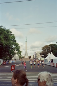 A group of people is participating in a running event on a city street, with a tall monument in the background. Tents and barriers line the street, and trees are visible on the left side. The weather appears partly cloudy.