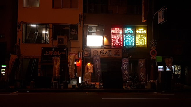 A nighttime urban scene showcasing a row of illuminated signs with Japanese text, attached to a multi-story building. The signs are vibrant in red, blue, green, yellow colors, highlighting various establishments and possibly bars or restaurants. Below the signs, banners and posters add to the lively street atmosphere, while the rest of the scene is dimly lit.