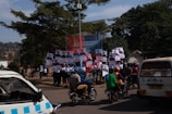 A group of people holding campaign posters stands on a sidewalk. Nearby, several motorcyclists drive by on the street. In the background, trees and a sign for public toilets are visible. A white van with text on the rear window is parked close by.