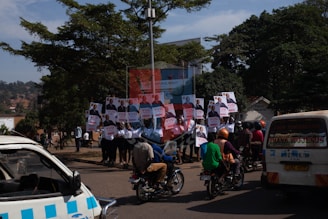 A group of people holding campaign posters stands on a sidewalk. Nearby, several motorcyclists drive by on the street. In the background, trees and a sign for public toilets are visible. A white van with text on the rear window is parked close by.