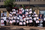 A group of people is standing outside a building, each holding up a large poster. The posters and banners feature an image of a person in a suit, along with text highlighting an event or promotion. Some participants are wearing matching white shirts, and they appear to be smiling or cheering. The setting is urban with some greenery, and the background features a modern, multi-story structure.