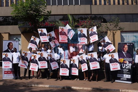 A group of people is standing outside a building, each holding up a large poster. The posters and banners feature an image of a person in a suit, along with text highlighting an event or promotion. Some participants are wearing matching white shirts, and they appear to be smiling or cheering. The setting is urban with some greenery, and the background features a modern, multi-story structure.