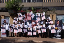 A group of people is standing outside a building, each holding up a large poster. The posters and banners feature an image of a person in a suit, along with text highlighting an event or promotion. Some participants are wearing matching white shirts, and they appear to be smiling or cheering. The setting is urban with some greenery, and the background features a modern, multi-story structure.