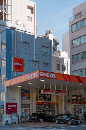 A gasoline station with a bright orange and red canopy featuring the 'ENEOS' logo. The station is surrounded by high-rise urban buildings under a clear blue sky. Several cars are parked at the station, and various signs in Japanese are visible.